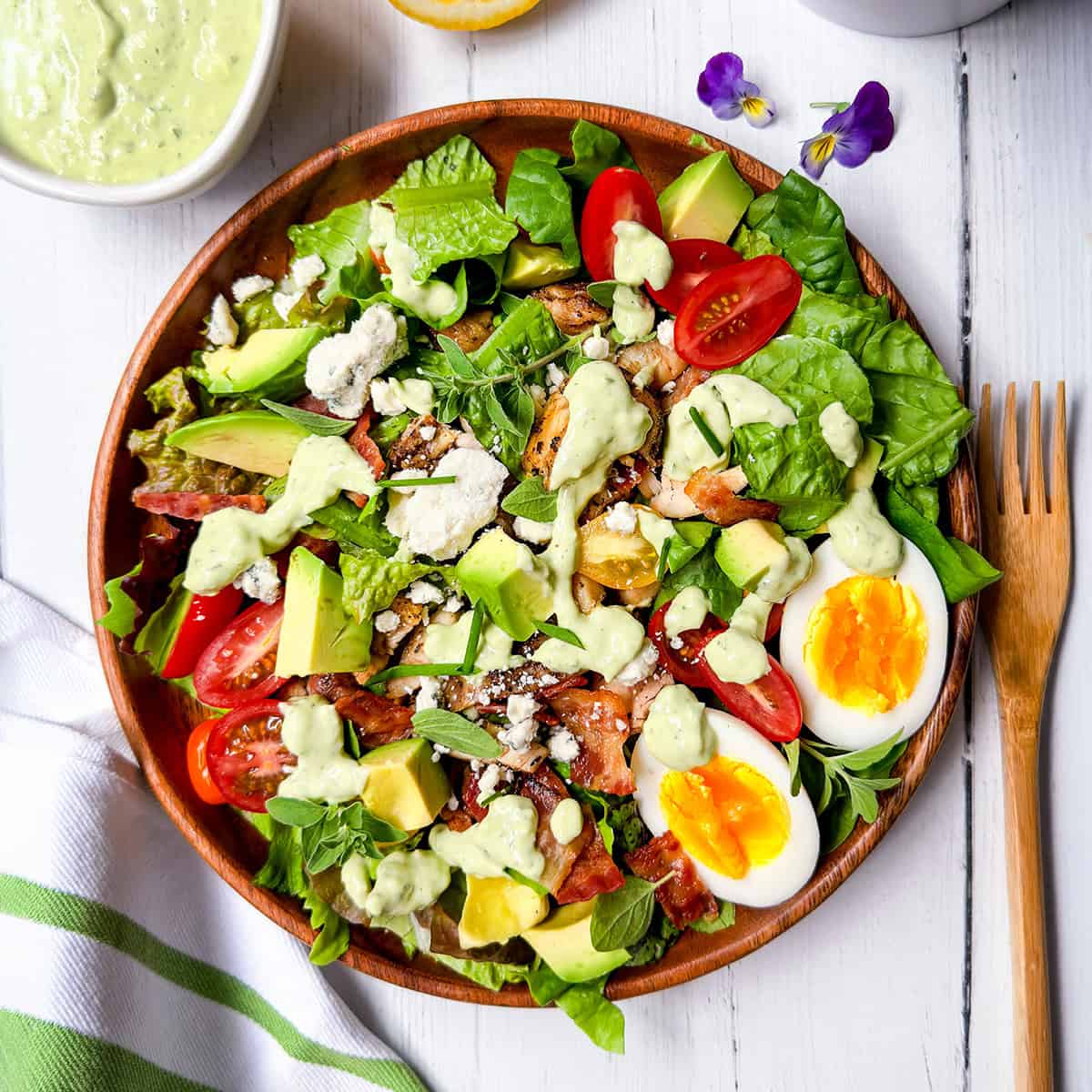 A wooden bowl filled with cobb salad-leafy greens, cherry tomatoes, avocado, bacon, feta cheese, a sliced boiled egg, and creamy dressing-sits on a white table with a fork and napkin beside it.