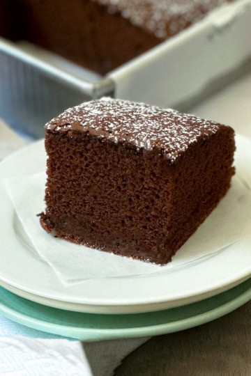 A single slice of decadent chocolate cake with powdered sugar on top sits on a small white plate, with a cake pan and more chocolate cake in the background.