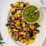 Sliced grilled ribeye steaks topped with corn kernels and green sauce, served on a white oval platter with a bowl of green chimichurri sauce and a spoon.