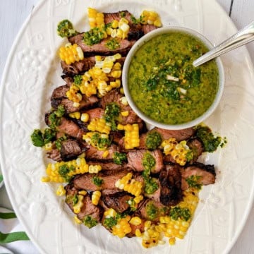 Sliced grilled ribeye steaks topped with corn kernels and green sauce, served on a white oval platter with a bowl of green chimichurri sauce and a spoon.