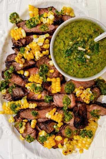 Sliced grilled ribeye steaks topped with corn kernels and green sauce, served on a white oval platter with a bowl of green chimichurri sauce and a spoon.