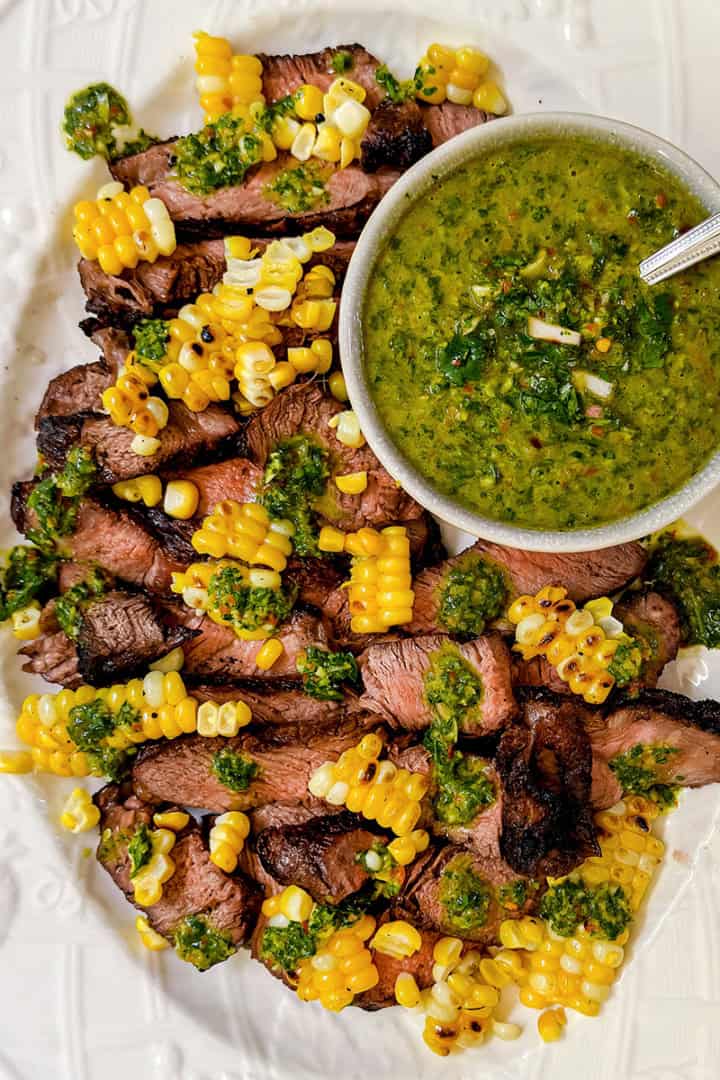 Sliced grilled ribeye steaks topped with corn kernels and green sauce, served on a white oval platter with a bowl of green chimichurri sauce and a spoon.