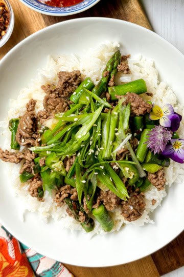 A bowl of white rice topped with Thai Ginger Beef, asparagus, sliced green onions, sesame seeds, and edible flowers, served on a wooden board.
