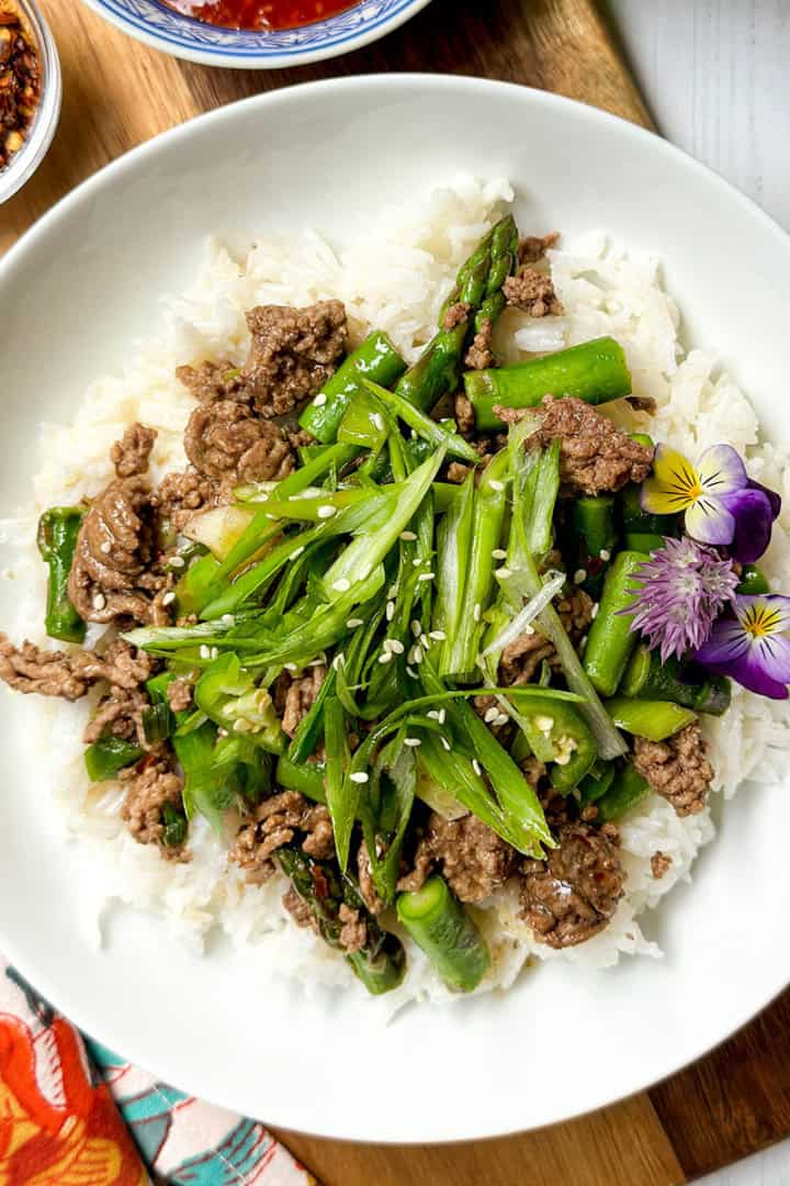 A bowl of white rice topped with Thai Ginger Beef, asparagus, sliced green onions, sesame seeds, and edible flowers, served on a wooden board.