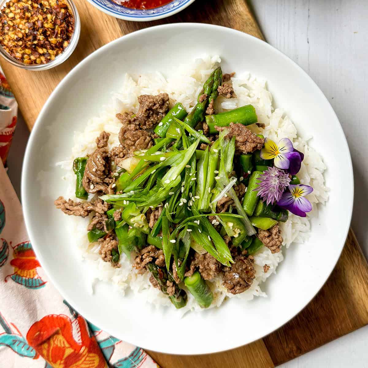 A bowl of white rice topped with Thai Ginger Beef, asparagus, sliced green onions, sesame seeds, and edible flowers, served on a wooden board.
