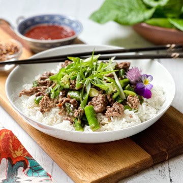 A bowl of white rice topped with Thai Ginger Beef, green beans, sliced scallions, and an edible flower, served with chopsticks and sauce on the side.