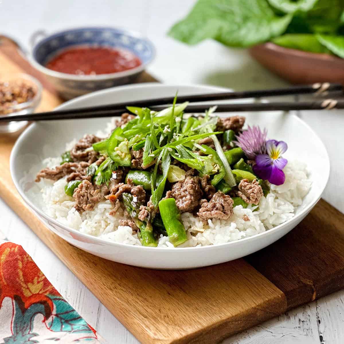 A bowl of white rice topped with Thai Ginger Beef, green beans, sliced scallions, and an edible flower, served with chopsticks and sauce on the side.