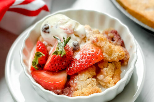 Top side view of a serving of strawberry cobbler in a white tart dish with melting vanilla ice cream