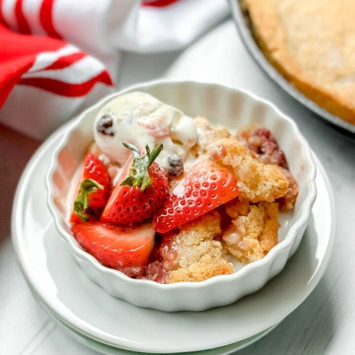 Top side view of a serving of strawberry cobbler in a white tart dish with melting vanilla ice cream