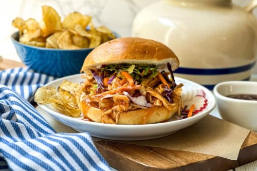 Side view of 3 ingredient Slow Cooker BBQ Chicken on a bun with coleslaw and chips