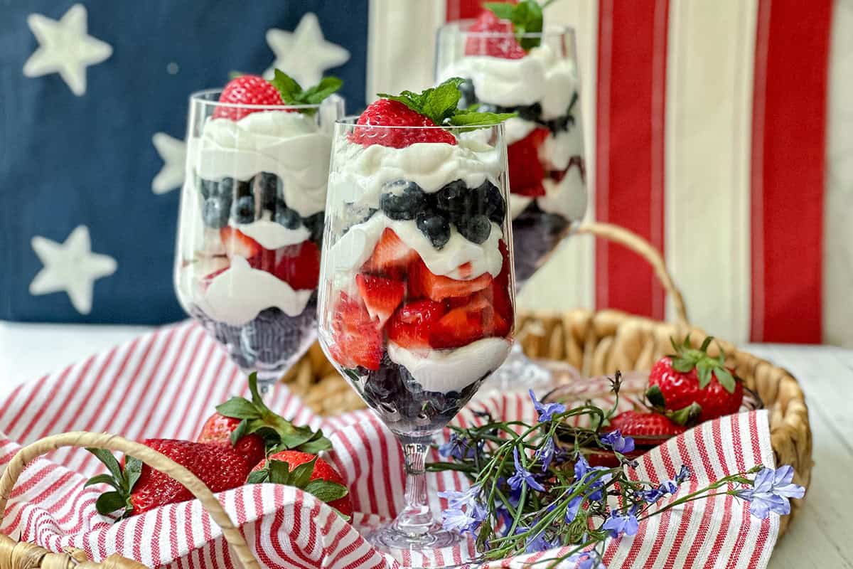 Three parfait glasses filled with layers of Greek yogurt, strawberries, and blueberries sit on a tray with a striped cloth, fresh strawberries, and small blue flowers. A classic American flag background highlights these festive parfait treats.
