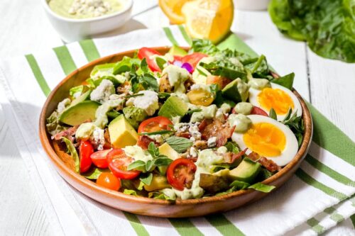 Closeup side view of Chicken Cobb Salad with lemon wedges, tomatoes, hard boiled eggs and avocado chunks in a wooden salad bowl