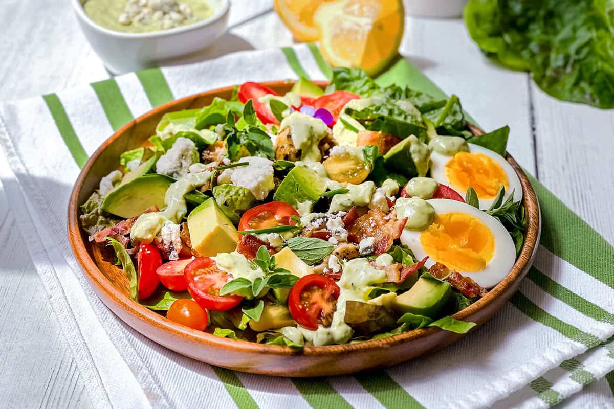 A wooden bowl holds a classic cobb salad with lettuce, avocado, cherry tomatoes, hard-boiled egg, chicken, bacon, cheese, and creamy dressing, set on a striped towel with lemon and extra dressing nearby.