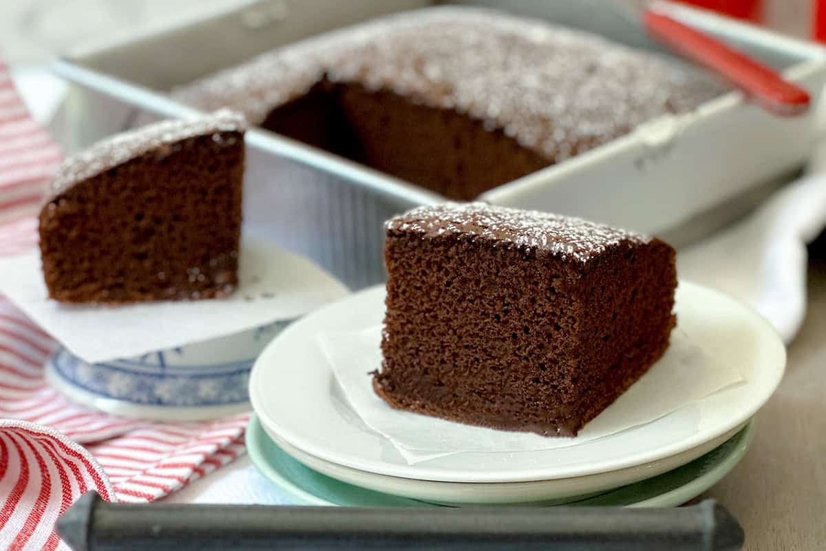 A square slice of rich chocolate cake sits on a white plate, with the rest of the chocolate cake and another slice tempting in the background.