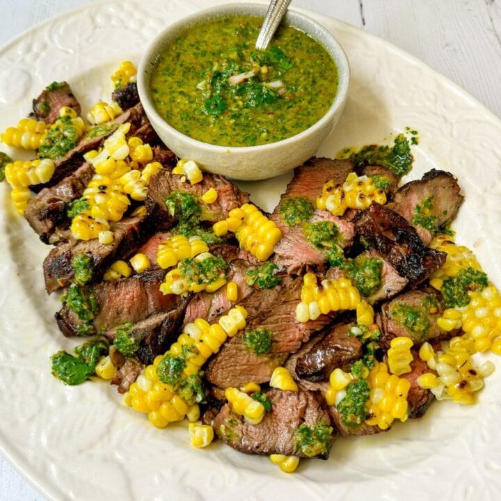 Side view of Grilled Ribeye Steaks, Chimichurri Sauce and grilled corn on a white oval platter