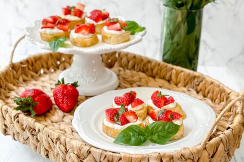 Side view of Strawberry Bruschetta on a white serving stand and a white scalloped plate in a woven tray