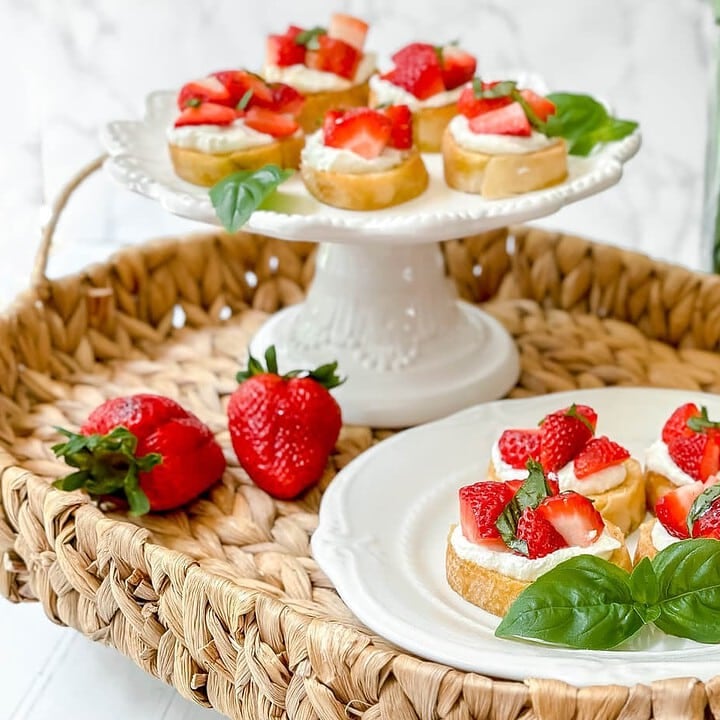 Side view of Strawberry Bruschetta on a white serving stand and a white scalloped plate in a woven tray