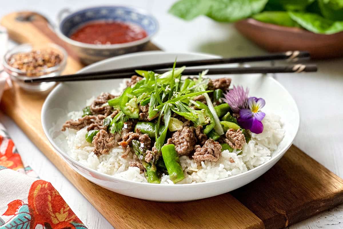 A bowl of white rice topped with Thai Ginger Beef, green vegetables, sliced scallions, and a purple edible flower, served with chopsticks on a wooden board.