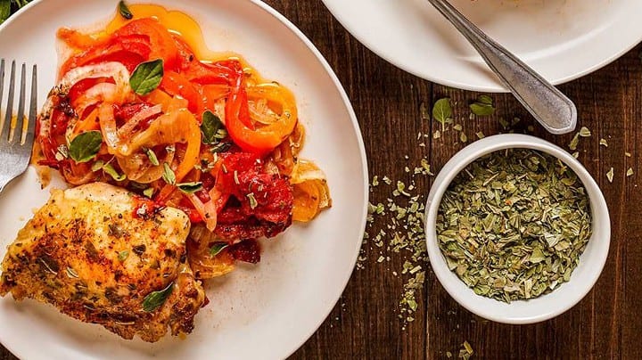 Baked chicken thighs served with roasted bell peppers, onions, and herbs on two plates&mdash;an ideal choice for August Mediterranean Dinners&mdash;accompanied by a baking tray and a bowl of dried herbs on a wooden table.