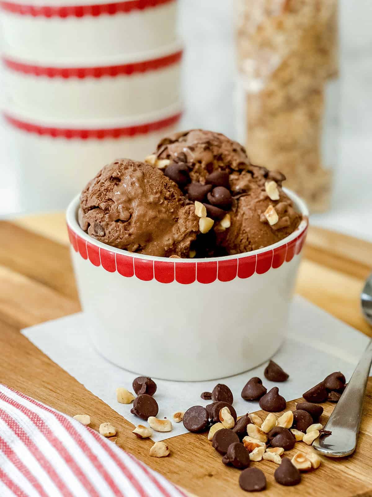 A bowl of chocolate protein ice cream topped with chocolate chips and chopped nuts sits on a wooden surface next to a spoon and a striped cloth.