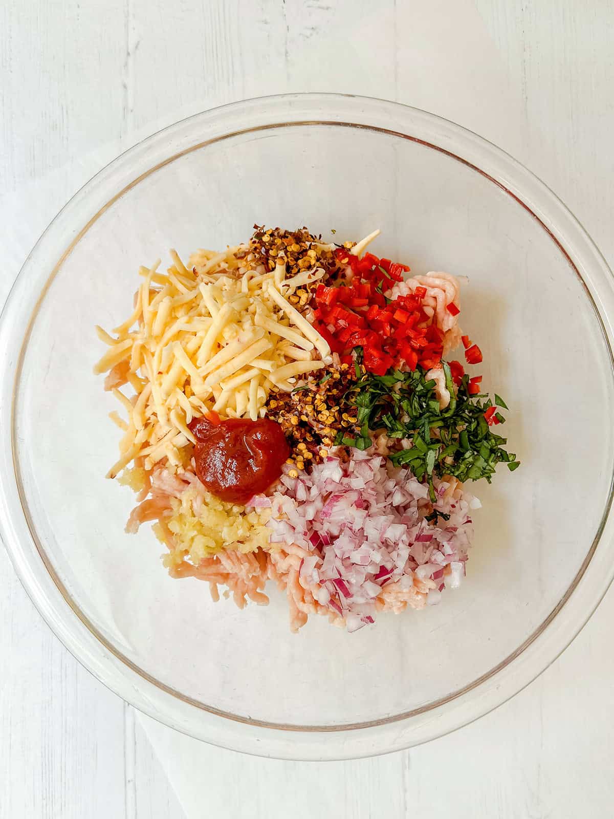 A clear glass bowl contains ingredients for firecracker chicken burgers: chopped red onions, shredded cheese, chopped herbs, diced red peppers, minced garlic, ground meat, seasonings, and a dollop of sauce arranged in separate piles.