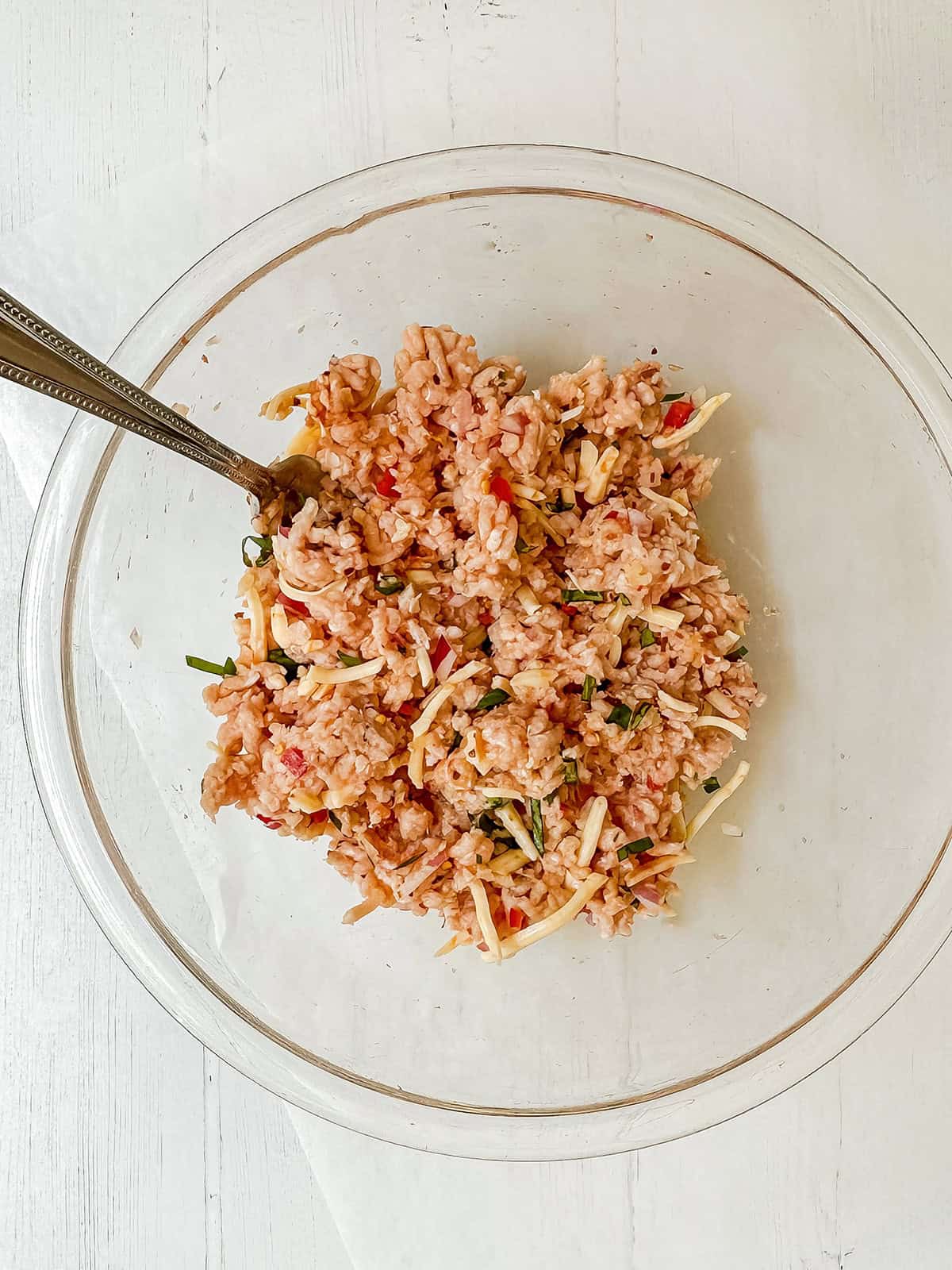 A clear glass bowl containing a ground chicken, shredded cheese, chopped vegetables, and herbs sits ready to complement spicy firecracker chicken burgers, with a metal spoon resting inside.