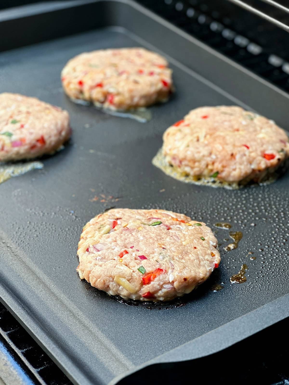 Four raw, uncooked firecracker chicken burger patties with visible chopped vegetables are cooking on a flat griddle.