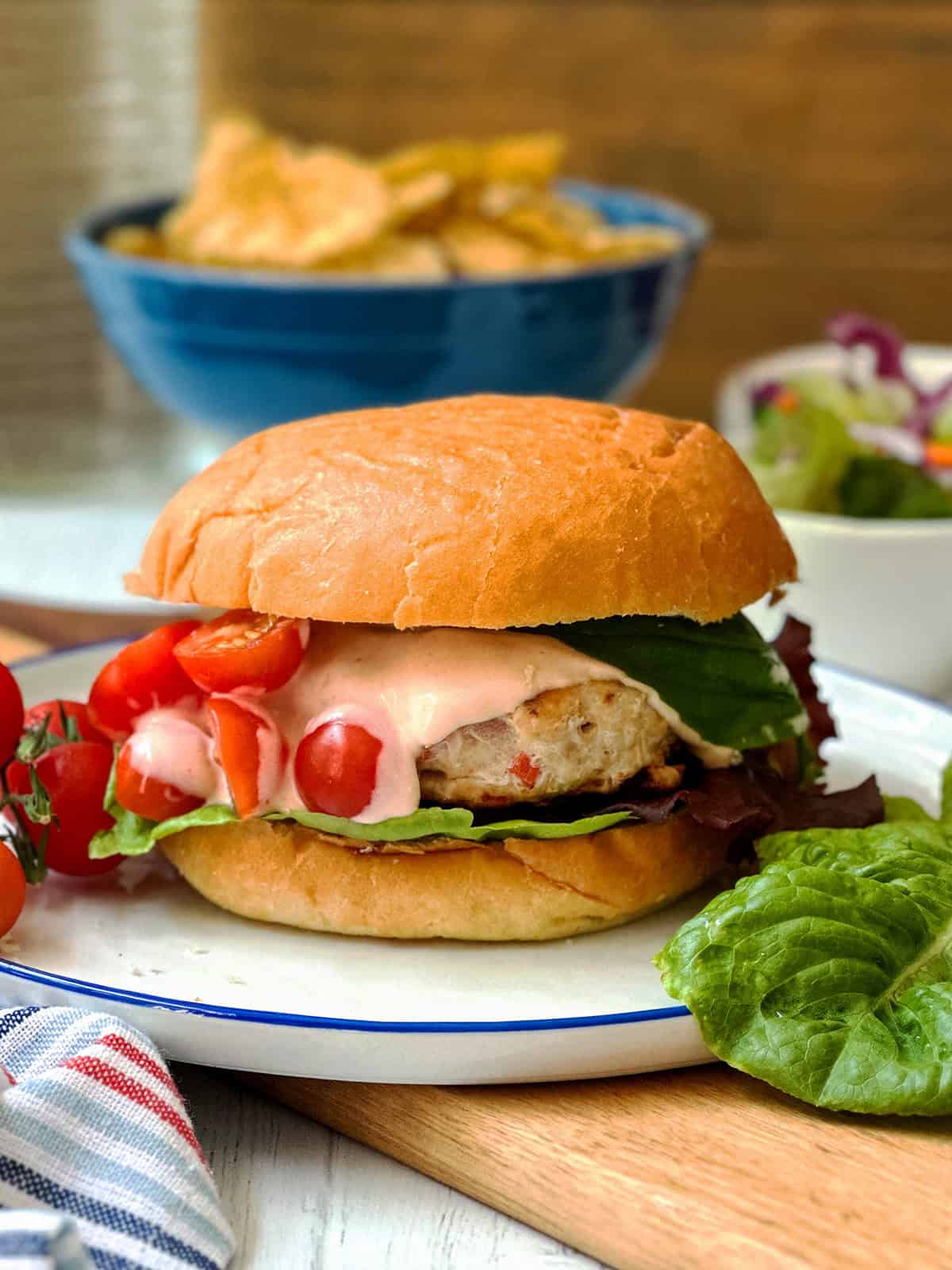 A firecracker chicken burger with lettuce, sliced cherry tomatoes, and pink sauce on a bun, served on a white plate with extra lettuce and cherry tomatoes on the side.