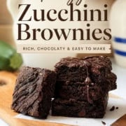 Three chocolate brownies, one drizzled with chocolate, are stacked on a wooden board. Text overlays highlight "Zucchini Brownies" as rich, chocolatey, and easy to make from 31Daily.com.