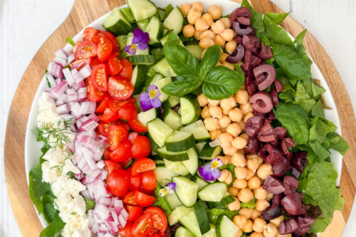 A round wooden board holds a vibrant Mediterranean Salad with rows of chopped lettuce, cucumbers, cherry tomatoes, red onions, chickpeas, kalamata olives, feta cheese, and a garnish of herbs and edible flowers.
