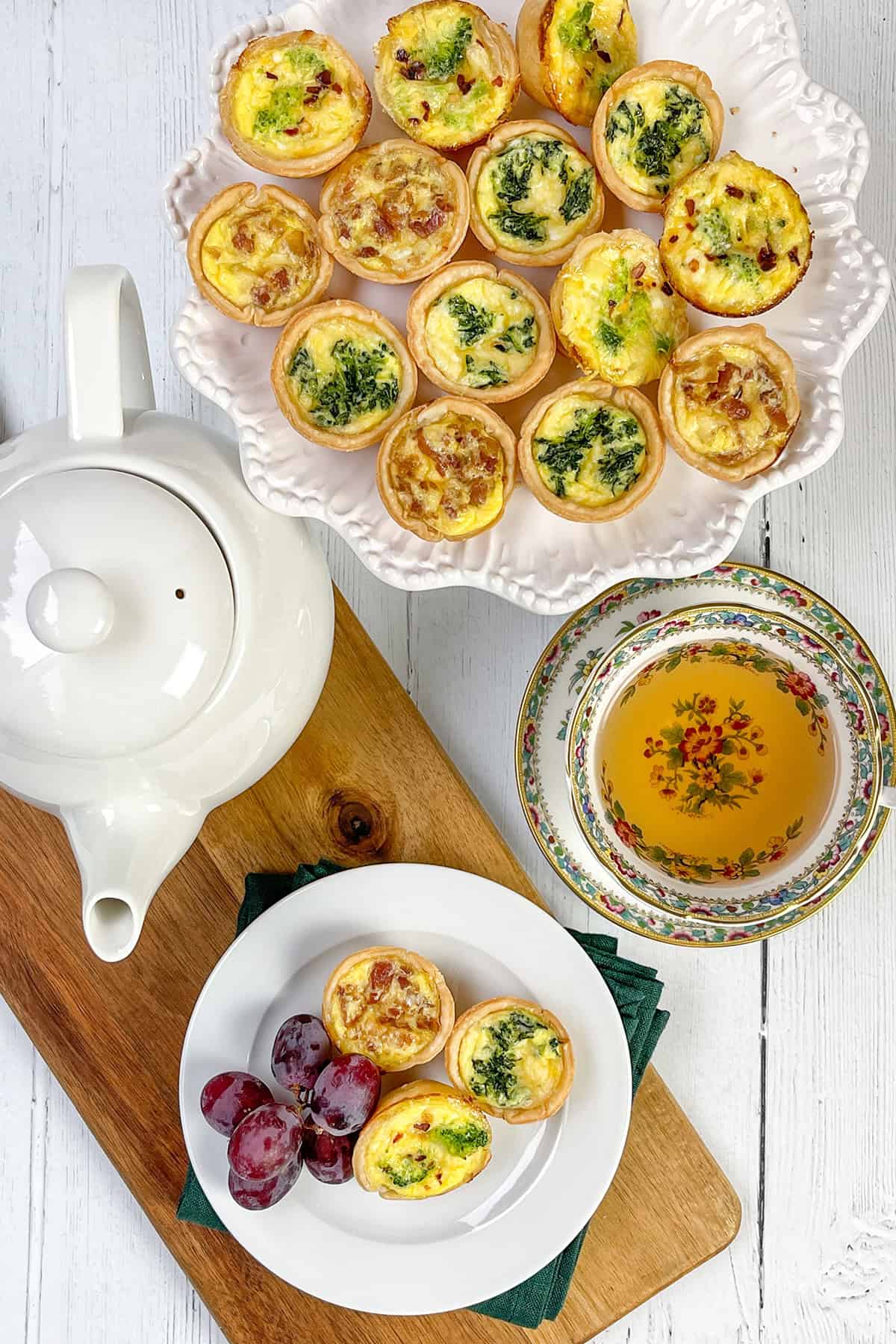 A plate of Mini Quiche, a teapot, a floral teacup filled with tea, and a small plate with Mini Quiche and grapes arranged on a wooden board atop a white table.