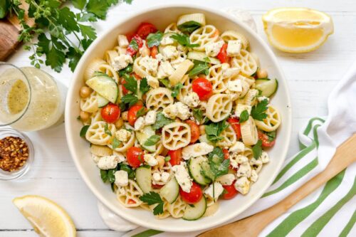 Top horizontal view of pasta salad with cucumbers, chickpeas, and tomatoes in a white bowl with fresh herbs in the background
