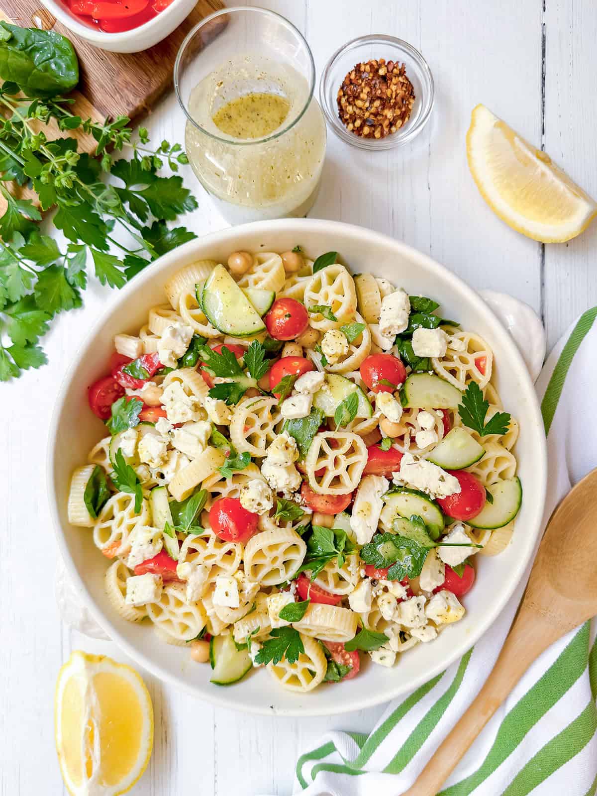 A bowl of pasta salad with cucumber, tomato, feta cheese, and parsley sits next to lemon wedges, a small bowl of red pepper flakes, and a jar of dressing on a white wooden table-an inviting spread perfect for trying out your favorite pasta salad recipe.