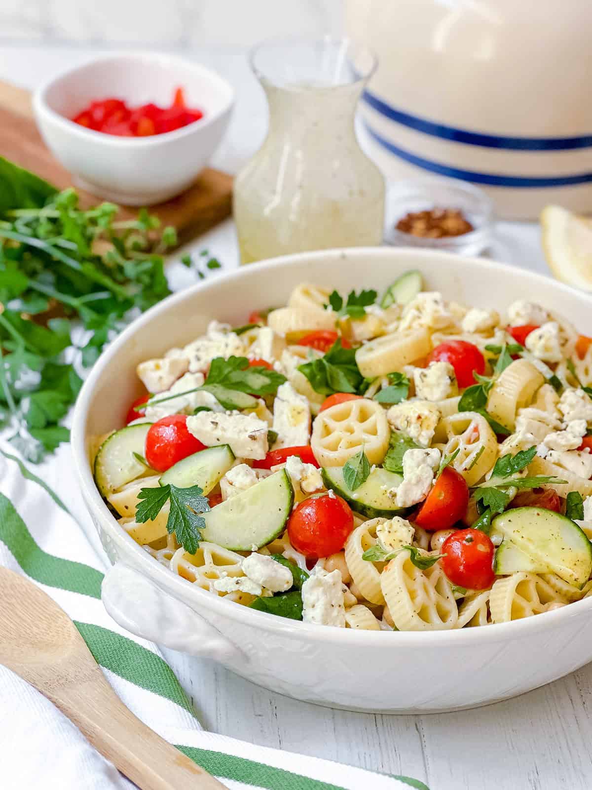 A bowl of pasta salad with cherry tomatoes, cucumber slices, feta cheese, and parsley, surrounded by a pitcher of dressing, herbs, and a bowl of chopped red peppers—this vibrant scene perfectly showcases a delicious pasta salad recipe.