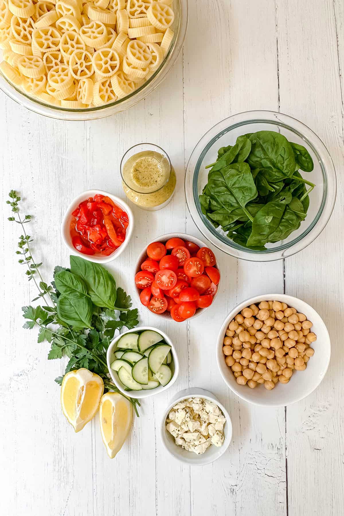 Bowls of dry pasta, spinach, chickpeas, cherry tomatoes, diced red peppers, cucumber slices, feta cheese, lemon wedges, herbs, and a glass of vinaigrette are arranged on a white table-perfect ingredients for creating a vibrant and fresh pasta salad recipe.
