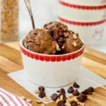 A white bowl with a red rim holds scoops of chocolate protein ice cream topped with chocolate chips and chopped nuts, with a spoon in the bowl.
