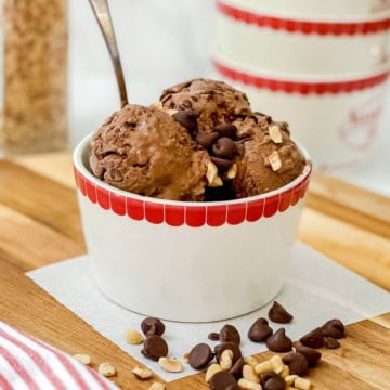 A white bowl with a red rim holds scoops of chocolate protein ice cream topped with chocolate chips and chopped nuts, with a spoon in the bowl.