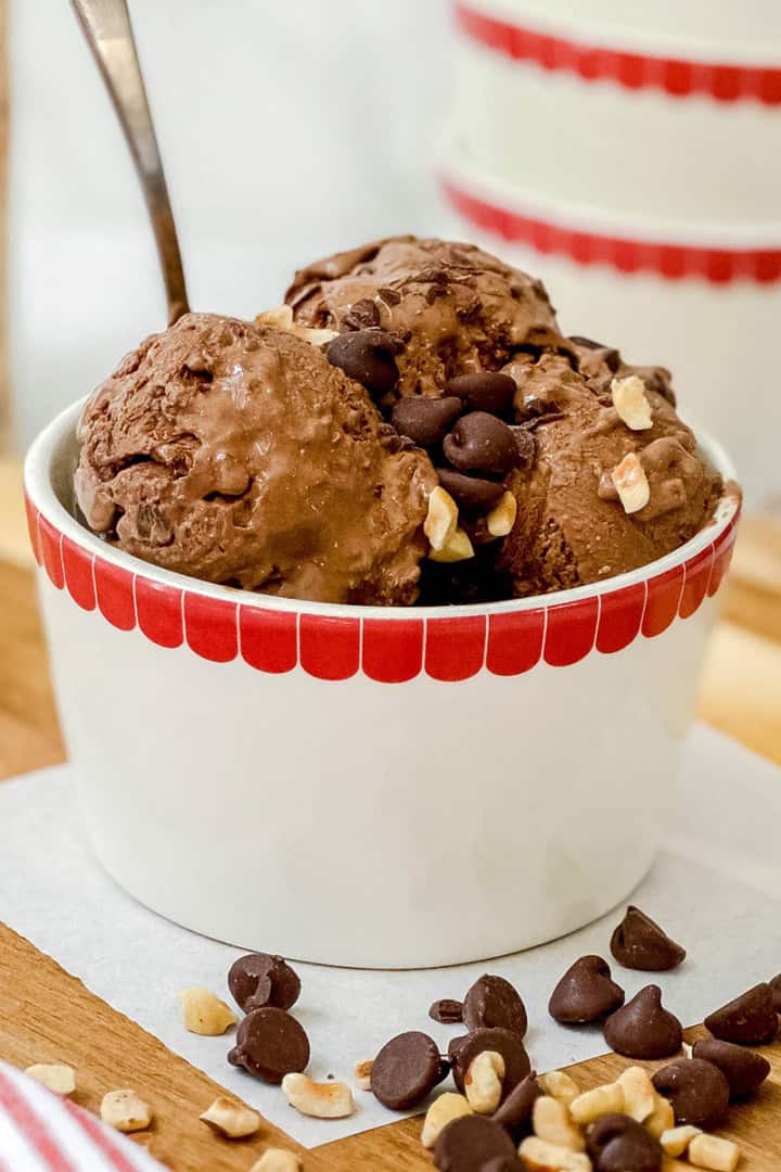 A white bowl with a red rim holds scoops of chocolate protein ice cream topped with chocolate chips and chopped nuts, with a spoon in the bowl.