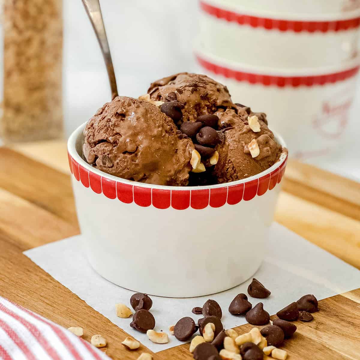 A white bowl with a red rim holds scoops of chocolate protein ice cream topped with chocolate chips and chopped nuts, with a spoon in the bowl.