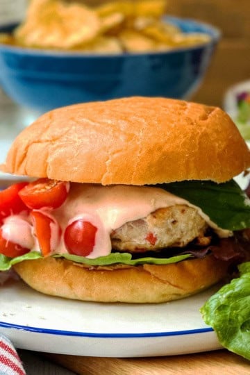 A firecracker chicken burger with lettuce, sliced tomatoes, and sauce on a bun, served on a plate with tomatoes and lettuce; chips and salad in the background.