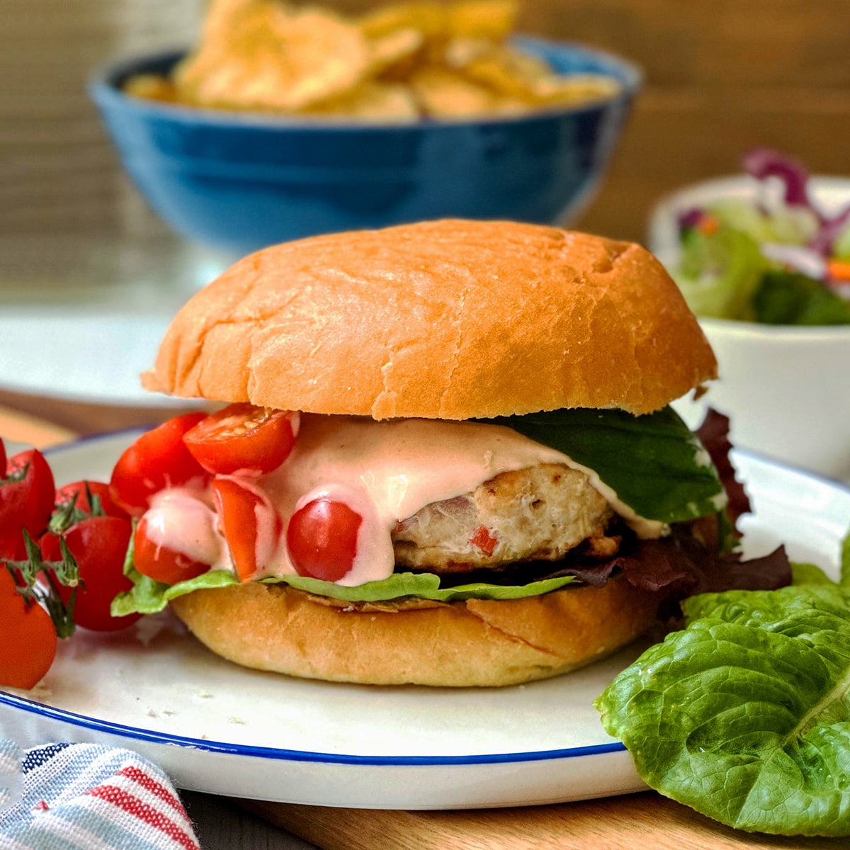 A firecracker chicken burger with lettuce, sliced tomatoes, and sauce on a bun, served on a plate with tomatoes and lettuce; chips and salad in the background.
