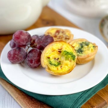 A white plate with three mini quiches and a small bunch of red grapes, set on a green napkin and wooden board. The golden, flaky mini quiche add an inviting touch to this elegant arrangement.