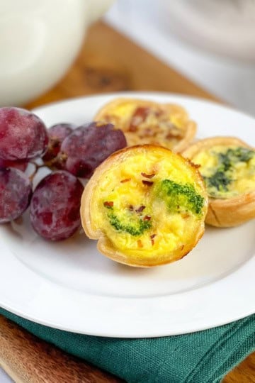 A white plate with three mini quiches and a small bunch of red grapes, set on a green napkin and wooden board. The golden, flaky mini quiche add an inviting touch to this elegant arrangement.