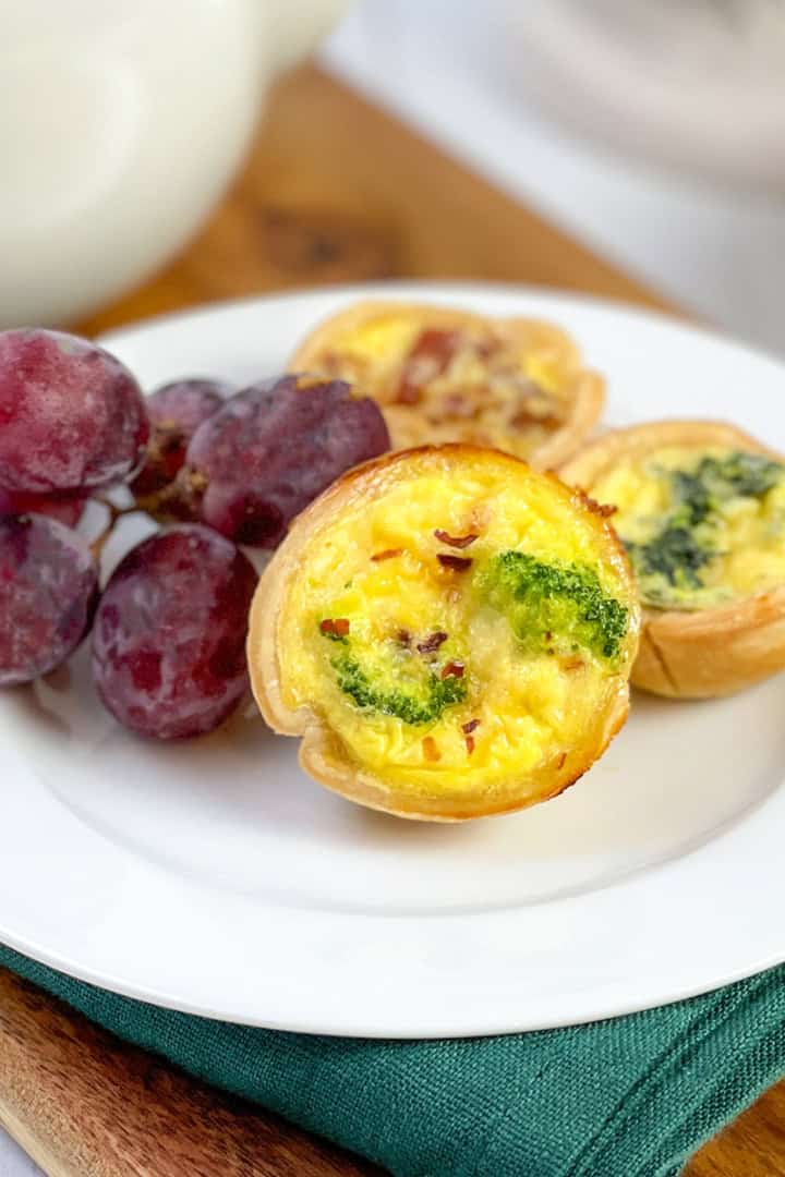 A white plate with three mini quiches and a small bunch of red grapes, set on a green napkin and wooden board. The golden, flaky mini quiche add an inviting touch to this elegant arrangement.