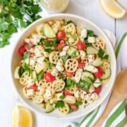 A bowl of pasta salad featuring wheel-shaped pasta, cherry tomatoes, cucumber slices, chickpeas, feta cheese, and parsley sits on a white table with lemon wedges and a wooden spoon nearby-a delicious pasta salad recipe perfect for summertime gatherings.
