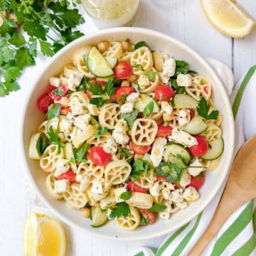 A bowl of pasta salad featuring wheel-shaped pasta, cherry tomatoes, cucumber slices, chickpeas, feta cheese, and parsley sits on a white table with lemon wedges and a wooden spoon nearby-a delicious pasta salad recipe perfect for summertime gatherings.