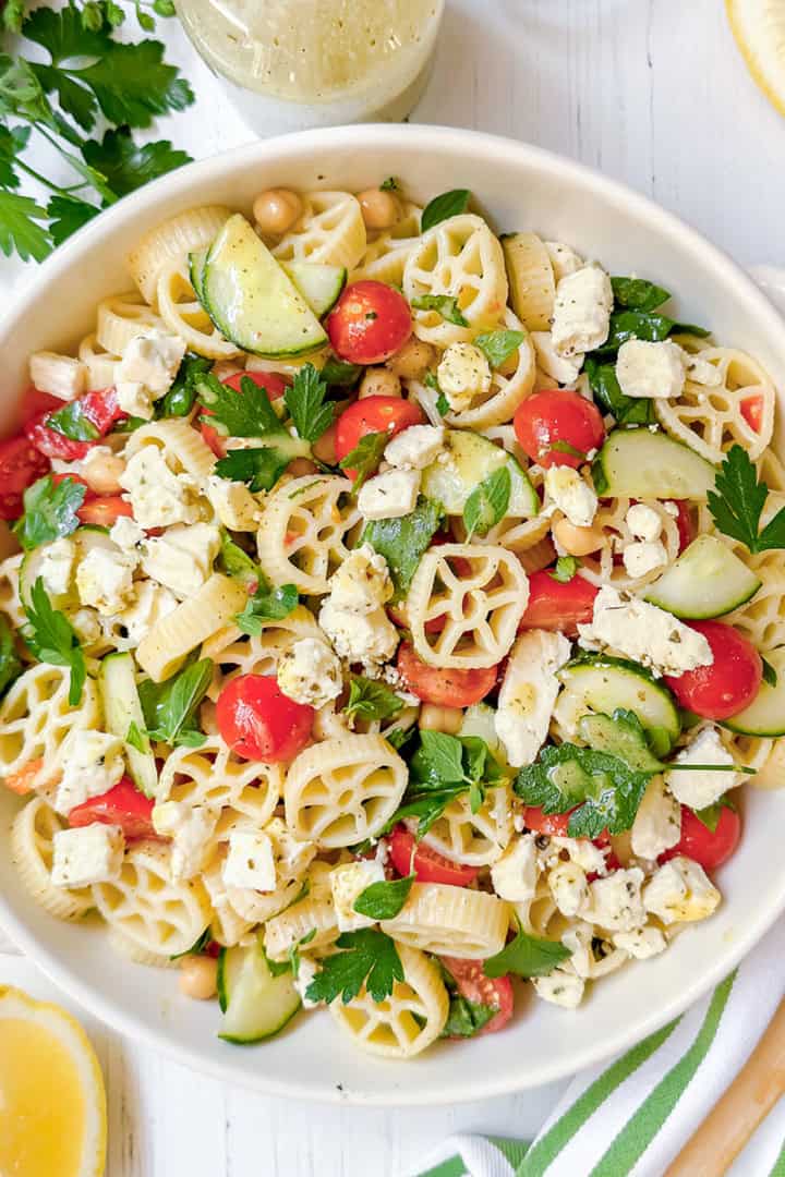 A bowl of pasta salad featuring wheel-shaped pasta, cherry tomatoes, cucumber slices, chickpeas, feta cheese, and parsley sits on a white table with lemon wedges and a wooden spoon nearby—a delicious pasta salad recipe perfect for summertime gatherings.
