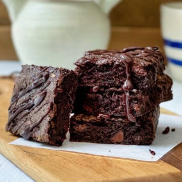 Three chocolate zucchini brownies stacked on parchment paper on a wooden board, drizzled with chocolate sauce, with a mug and pitcher in the background.