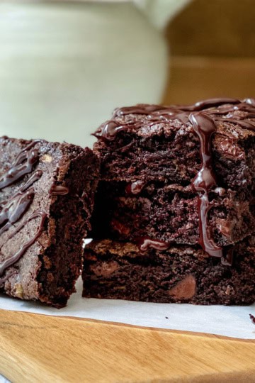 Three chocolate zucchini brownies stacked on parchment paper on a wooden board, drizzled with chocolate sauce, with a mug and pitcher in the background.