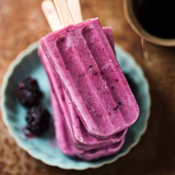 Three purple blackberry Greek yogurt popsicles stacked on a turquoise plate with two blackberries beside them, shown from above.